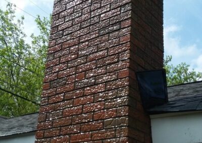 Tall red brick chimney against blue sky with trees and residential roofing visible
