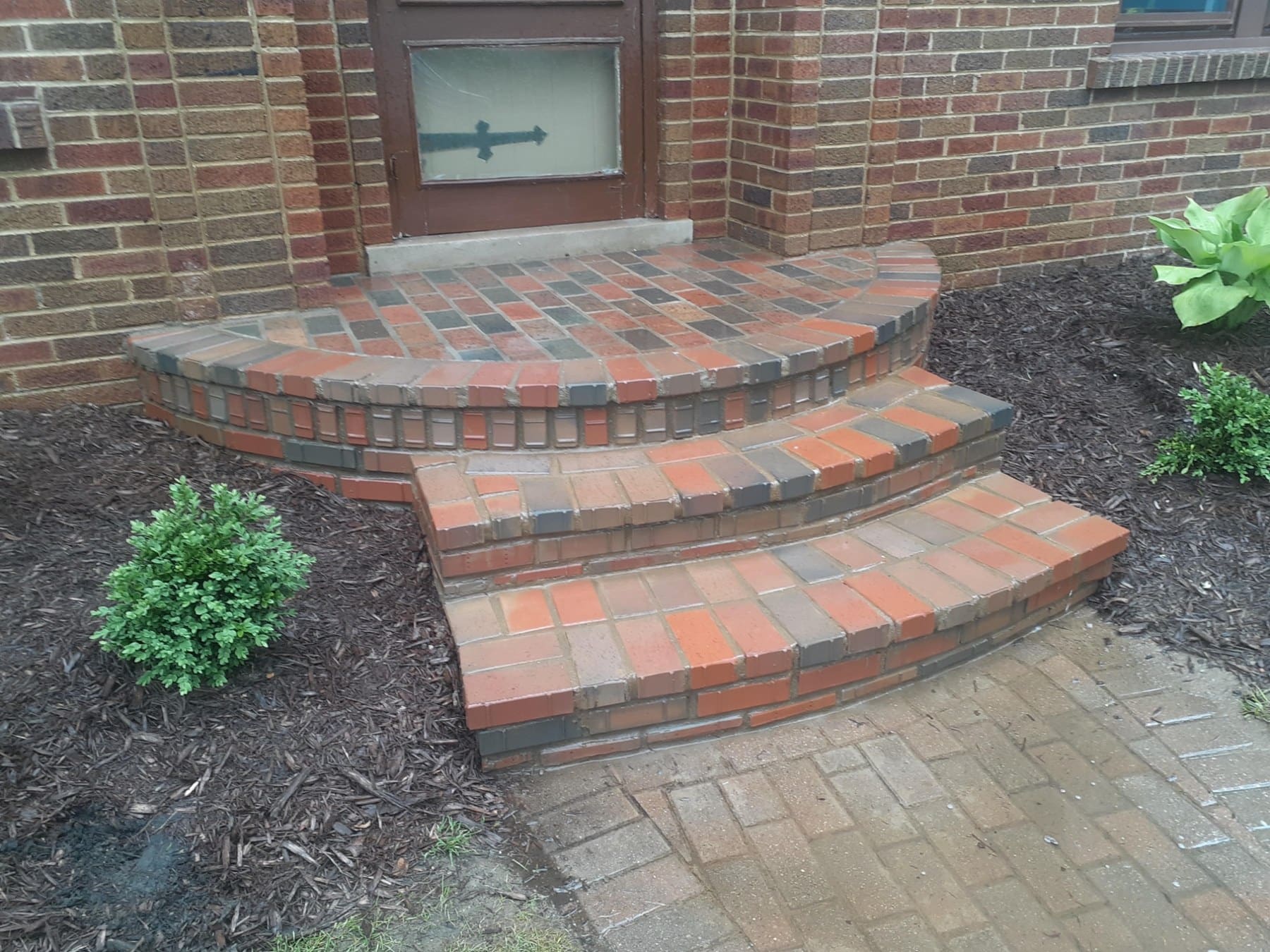 Curved brick entryway with three steps leading to a brown door with directional arrow window, flanked by landscaping and dark mulch