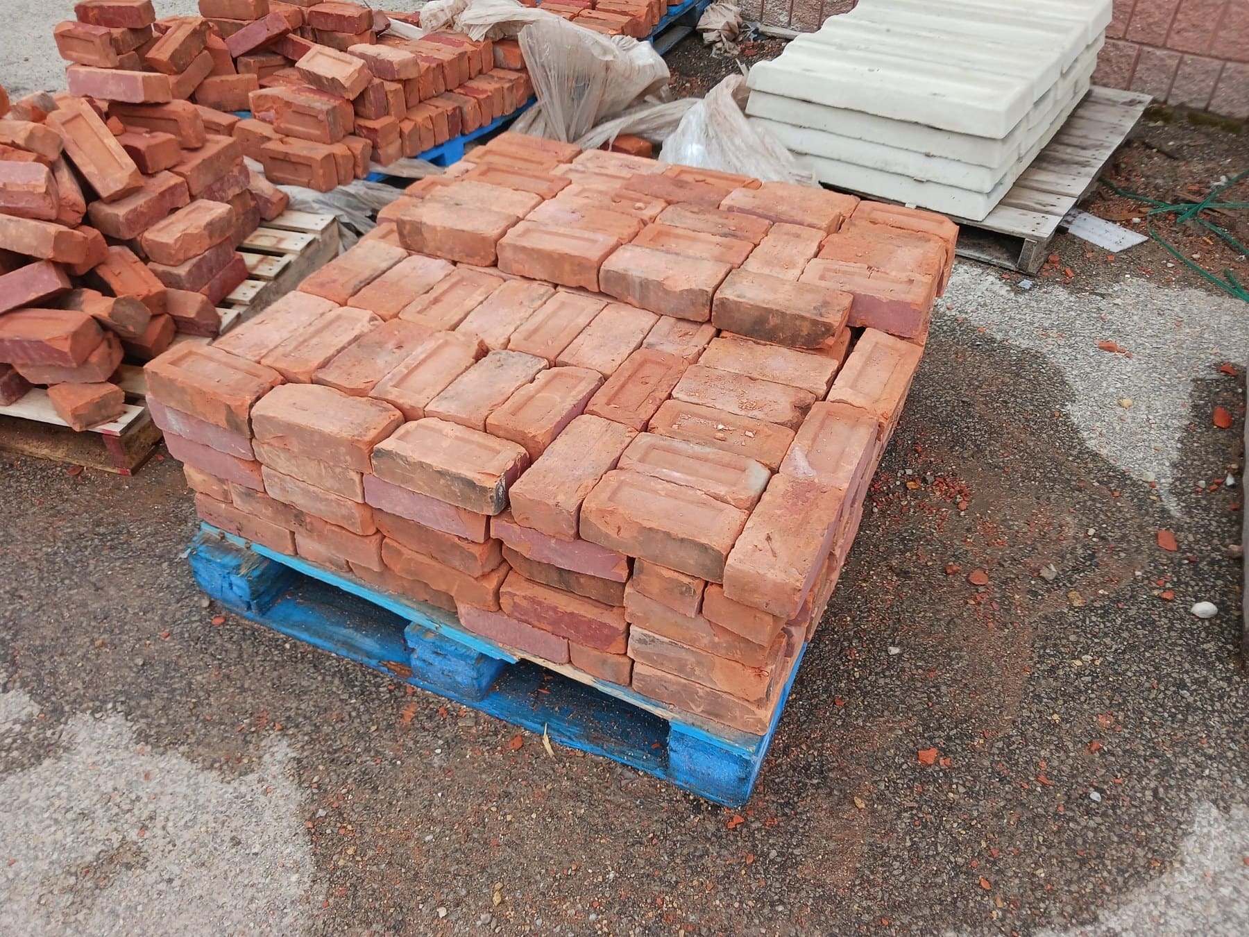 Pallet of stacked red clay bricks at construction materials yard with additional bricks and white pavers visible in background