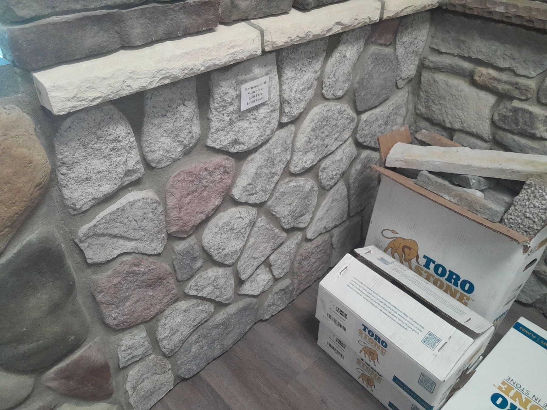 Stone veneer wall display with product boxes labeled Toro stacked on floor beside sample masonry materials