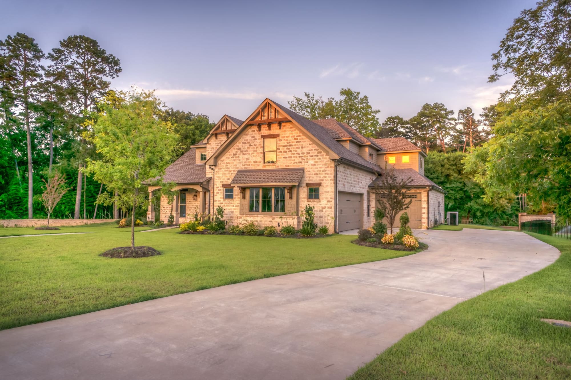 Luxury stone home with gabled roof and three-car garage surrounded by mature trees at dusk