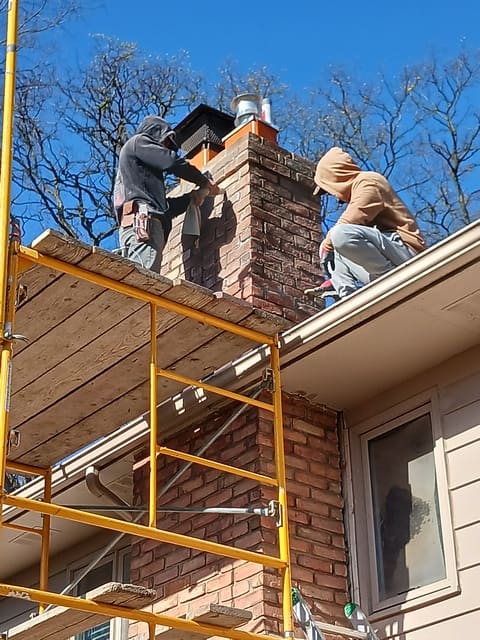 Two workers on yellow scaffolding repairing brick chimney against clear blue sky