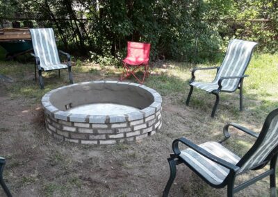 Stone fire pit surrounded by four lawn chairs in backyard setting with trees